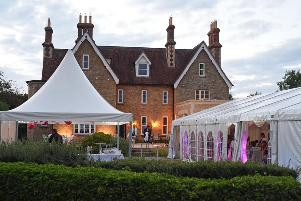 Large house with white tents set up in a garden; event in progress with lights on.