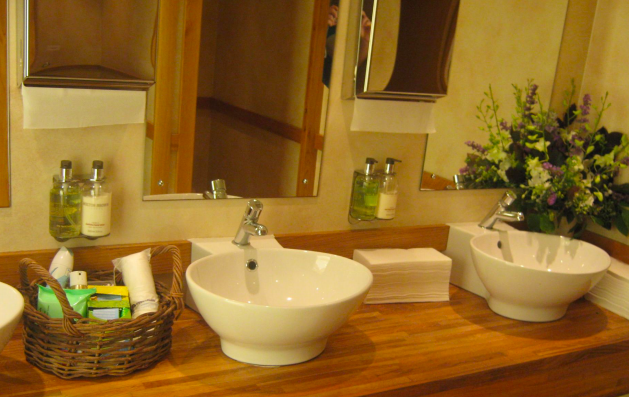 Bathroom with two sinks, mirrors, and paper towel dispensers.  Wooden countertop, toiletries in basket, floral arrangement.