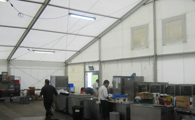 Inside a large white tent, several people work around stainless steel kitchen equipment.