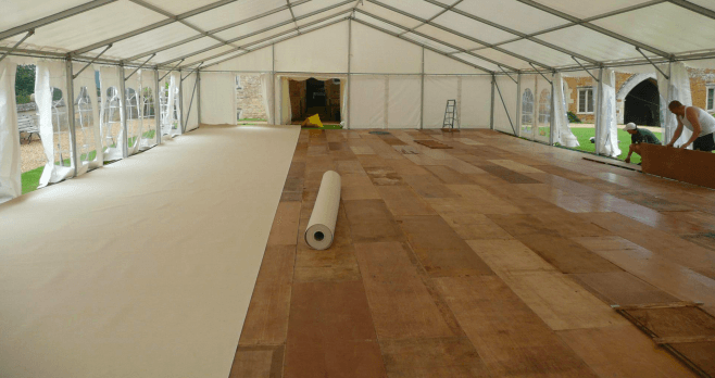 Interior of a tent with a wooden floor. A roll of white flooring material lies near the center. Man works at the tent's edge.