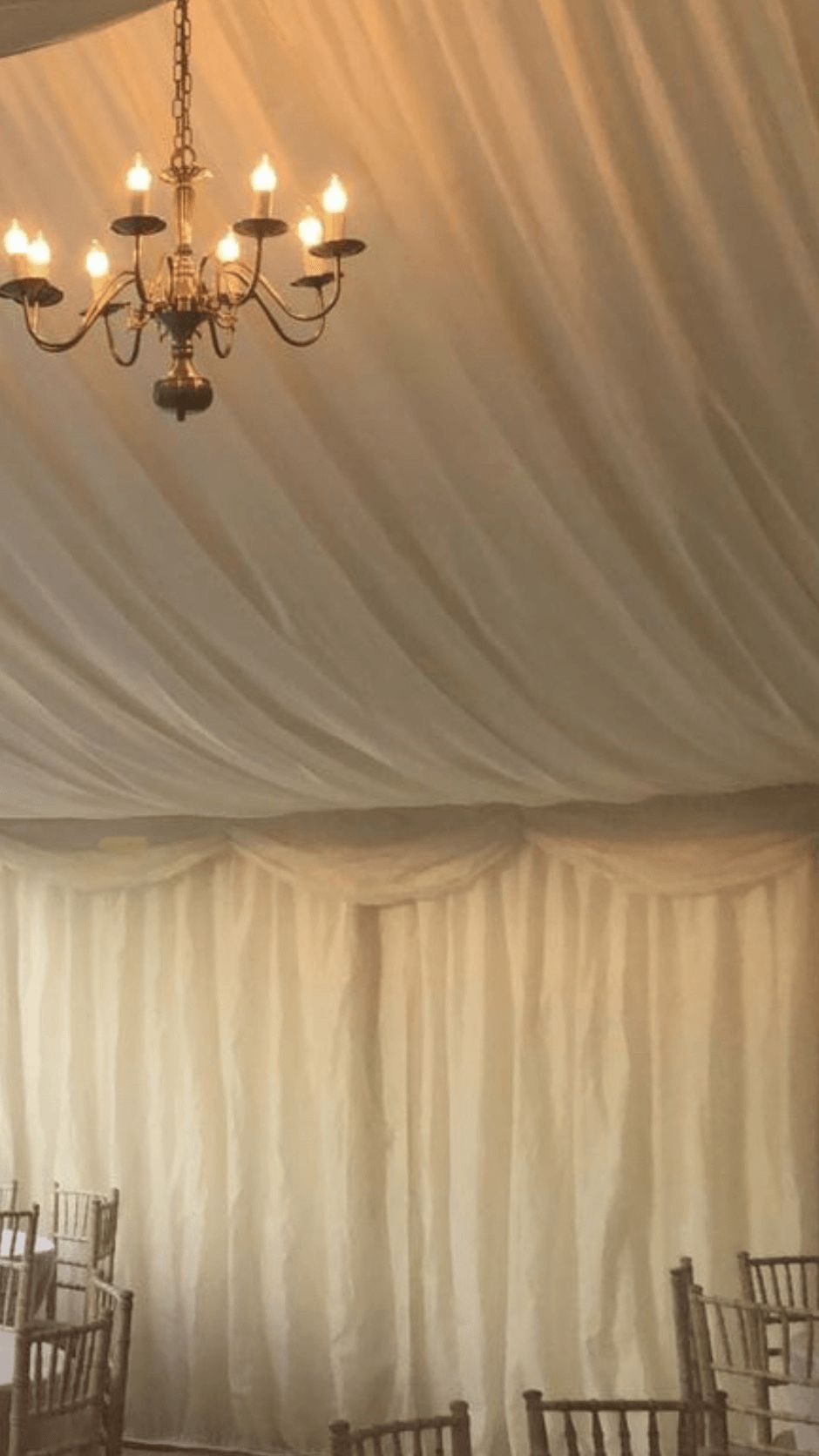 A tent interior with draped white fabric ceiling and walls. A lit chandelier hangs from above.