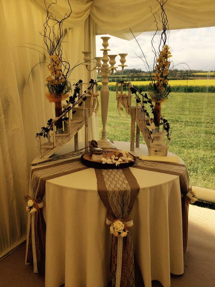 Table display with wedding decorations: floral arrangements, candelabra, and burlap runners inside a tent, with a field view.