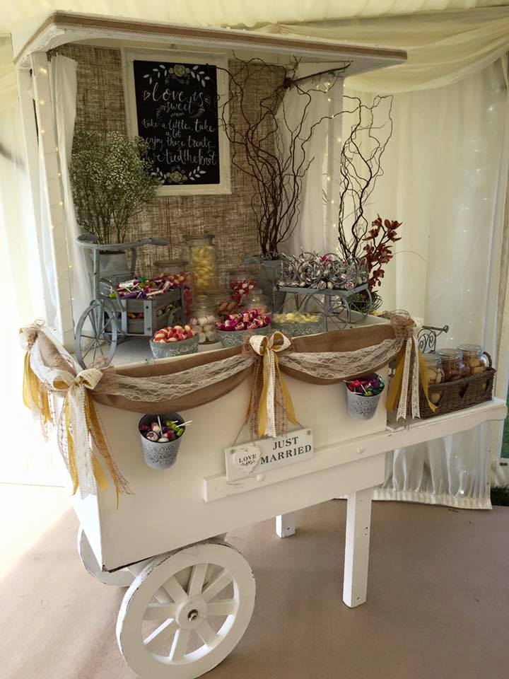White candy cart decorated for a wedding, with jars of sweets, burlap, and floral accents.