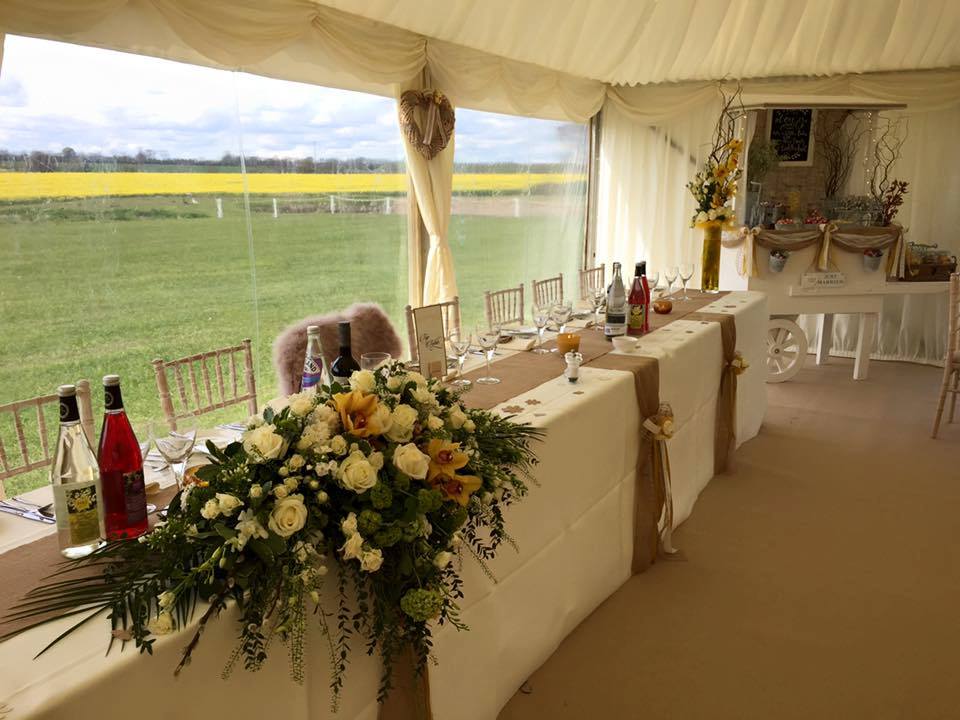 Wedding reception tent with table setting, floral arrangements, and scenic view of green field and yellow flowers.