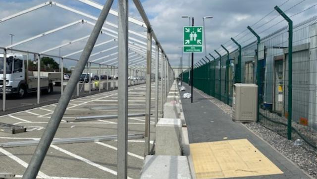 Outdoor area with vehicles and tent. Fenced perimeter with emergency signage, gray and green tones.