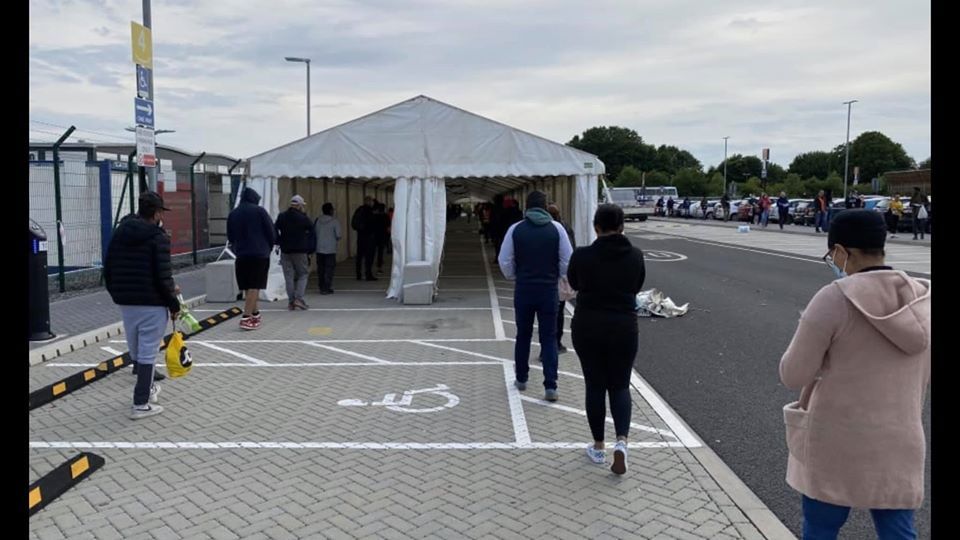 People queue at a tent entrance in a parking lot. White tent, concrete paving, overcast sky.
