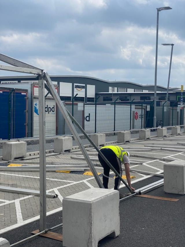 Person in a yellow vest assembling metal framework in a parking lot, with DPD signage in the background.