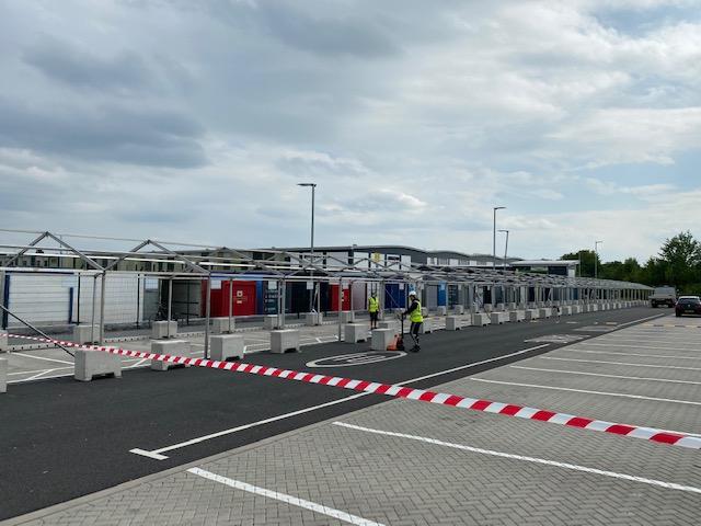 Outdoor testing site with multiple enclosed booths, barricades, and workers. Cloudy sky overhead.