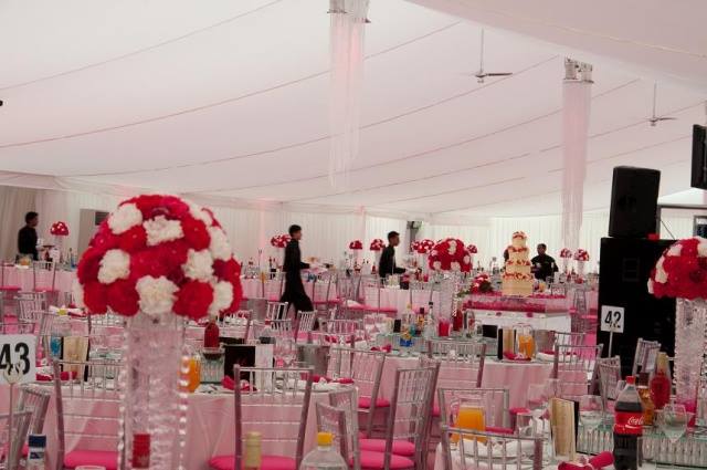 Inside a large, decorated tent, tables are set for an event with red and white floral centerpieces and pink tablecloths.