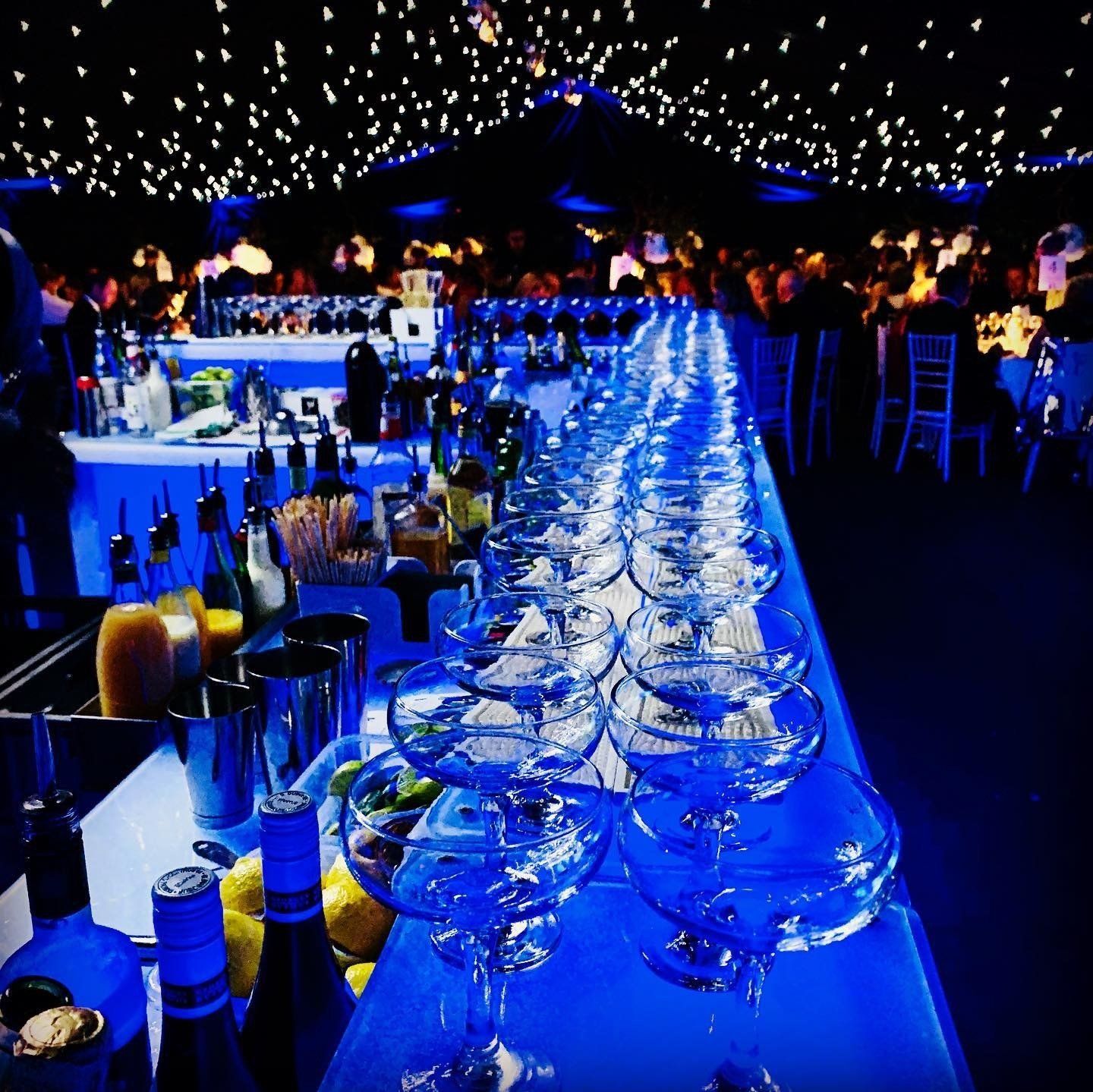 Long, blue-lit bar with glasses, bottles, and bartender, in a dimly lit event space with tables and string lights.
