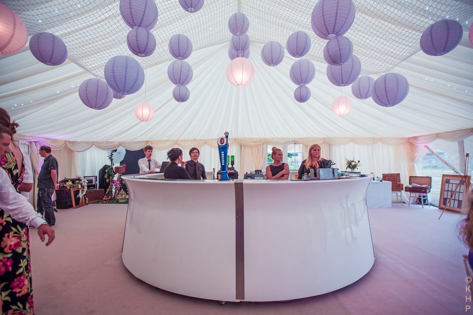 White bar inside a tent, decorated with purple paper lanterns, people are serving drinks.