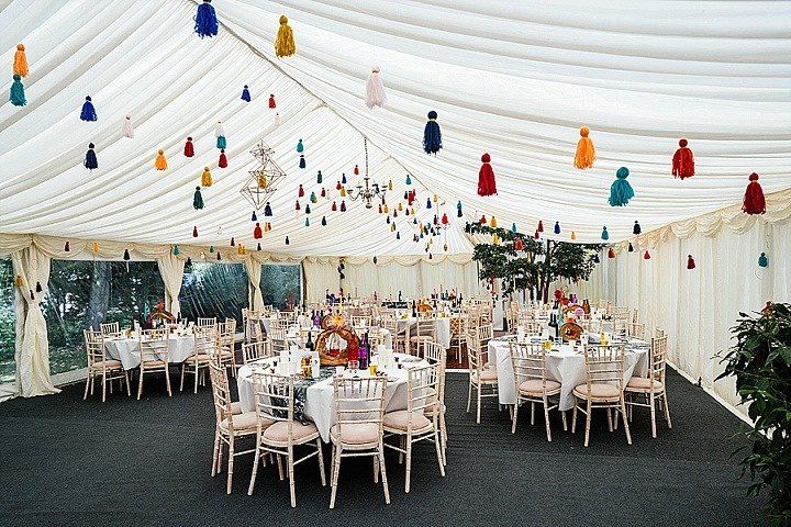 Wedding reception tent with decorated tables, tassels hanging from the ceiling, and windows in the background.