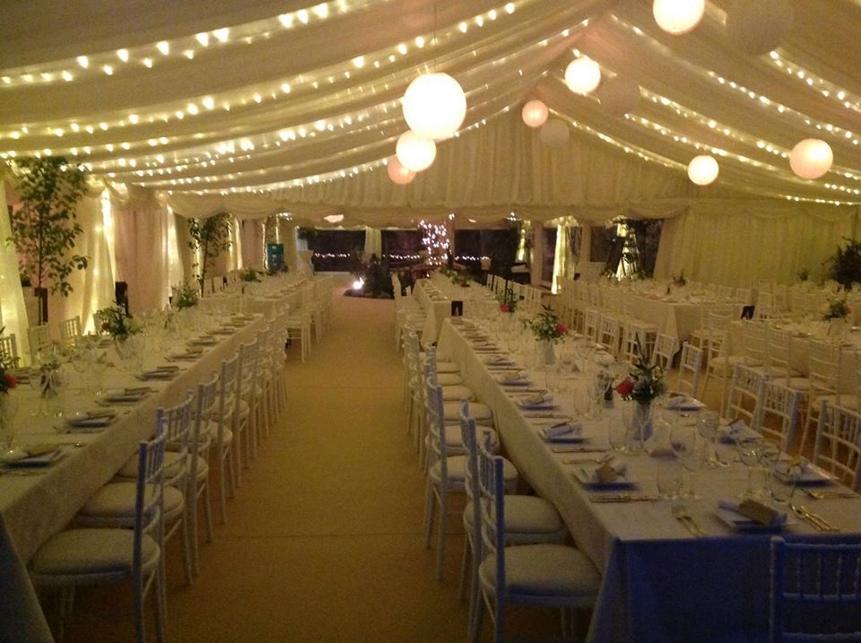 Wedding reception inside a tent, long tables set with white linens and chairs, lit by string lights and paper lanterns.
