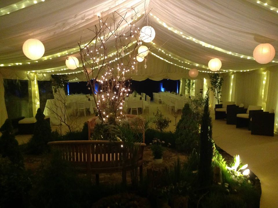 A tent interior decorated with string lights, paper lanterns, and a garden setting, with seating.