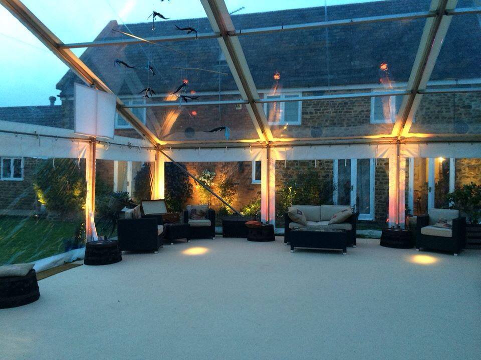 Clear tent with seating, lit with warm lights, set outdoors with a house in the background.