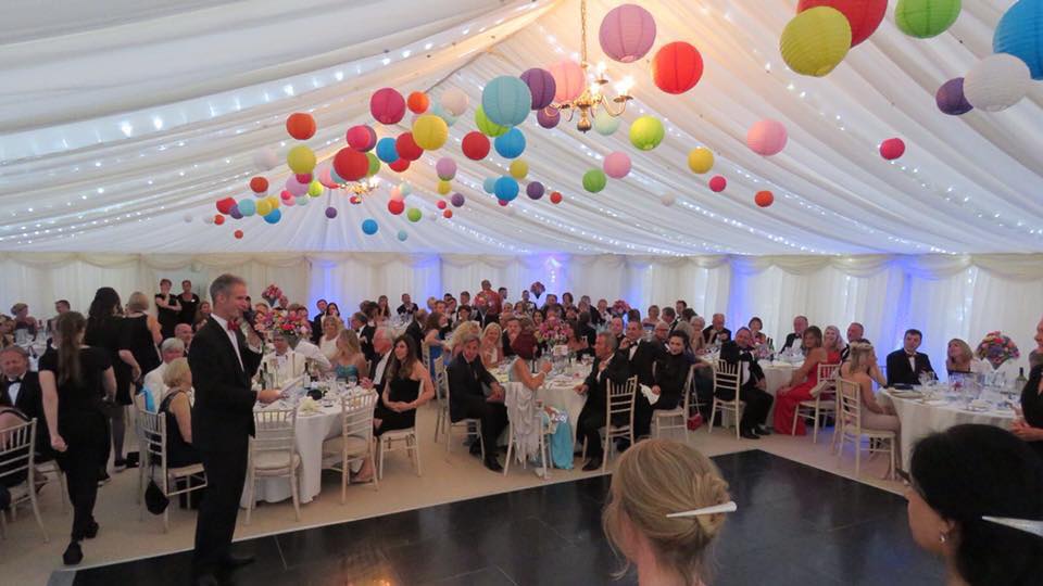 Large party in a tent; people seated at tables, speaker addressing crowd, colorful lanterns, and white draping.