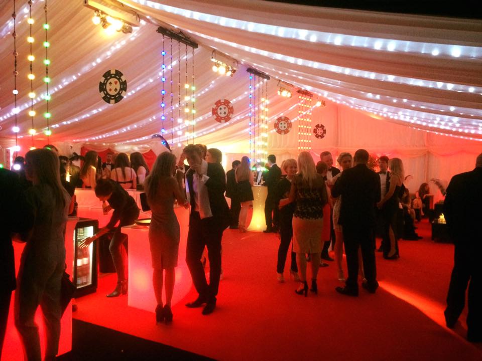 People in formal attire at a casino-themed event under a white tent with red carpet and poker chip decor.