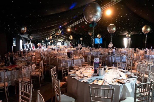 Tables set for an event inside a dark tent, silver balloons overhead, with many chairs around tables.