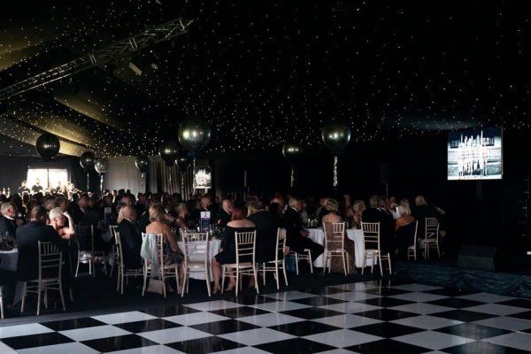 Black-and-white gala with people seated at tables, checkered dance floor, black balloons, and starry ceiling.