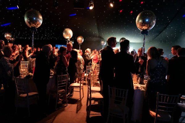 People in formal attire at a gala, applauding around tables with silver balloons overhead.