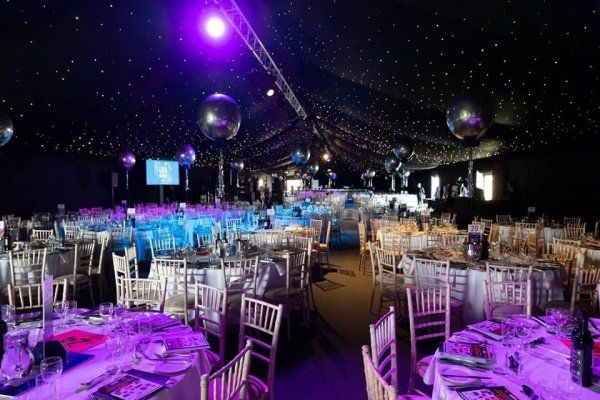 An event tent with tables set for a party, featuring blue and purple lighting, disco balls, and a starry ceiling.