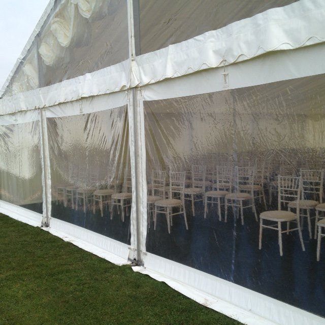 Clear-walled tent with rows of white chairs inside, set on a dark floor over green grass.