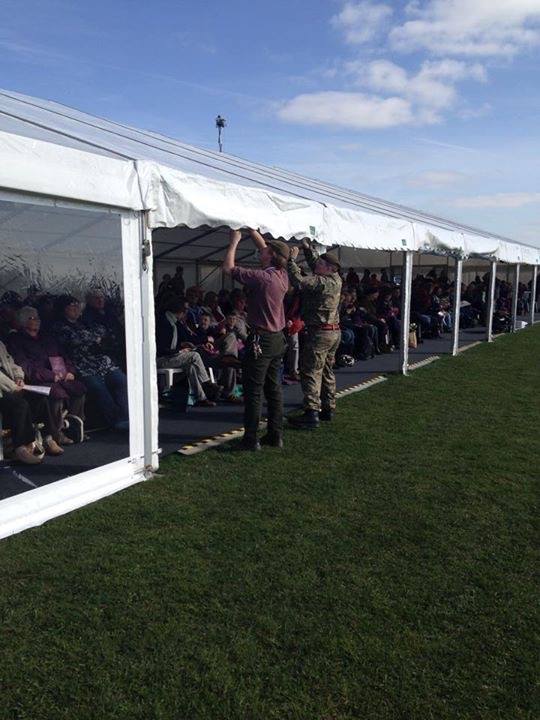 People setting up a long white tent on a grassy field; others sit inside.