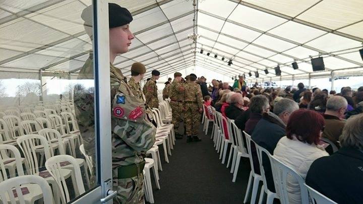 Soldiers stand in formation near the entrance of a tent where an event is taking place. Attendees seated.