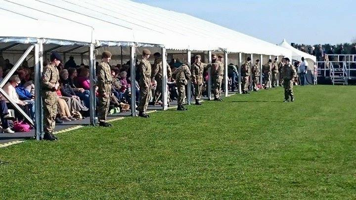 Soldiers in uniform stand in a line on grass, next to a white tent; spectators seated nearby.