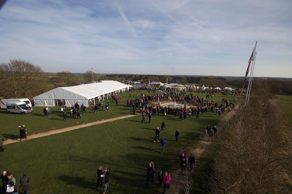 Large crowd gathered on a grassy field near a large white tent. Flags fly in the distance.