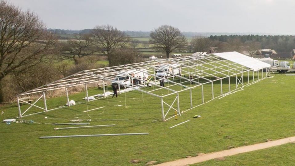 A partially built white tent frame in a field with vehicles and workers. Overcast day.