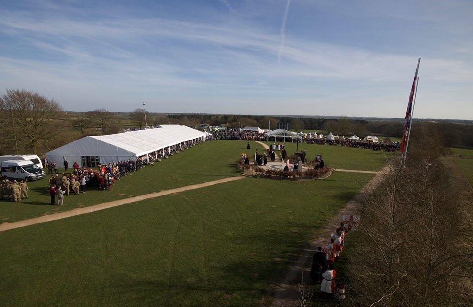 Large outdoor event with white tents, people, flag, and green field under a blue sky.