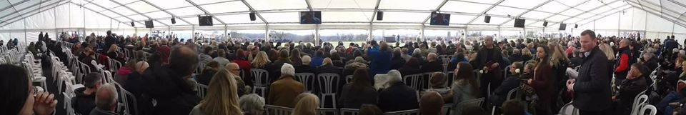 Large crowd seated in a tent. Many people are in attendance with chairs and tables set up.