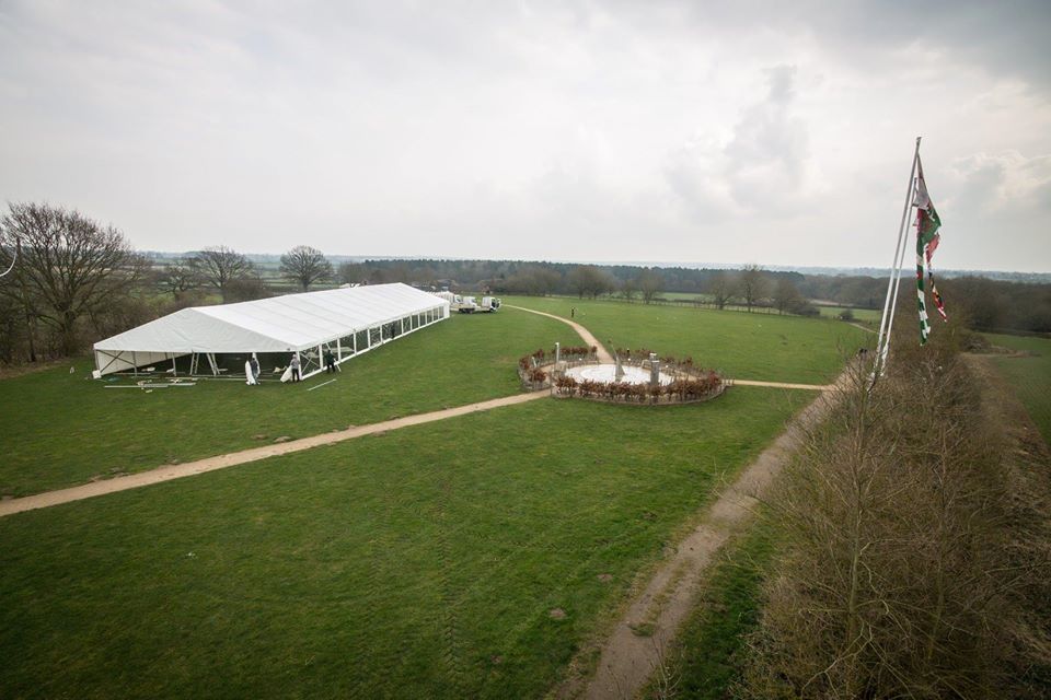 White tent and circular arrangement on a grassy field; flags fly nearby under a cloudy sky.