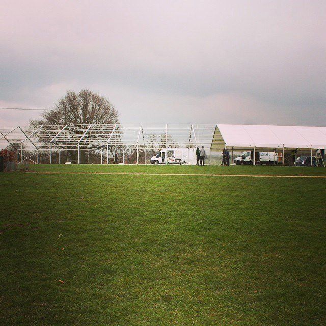 A large white tent under construction in a green field with a cloudy sky. Workers are present.