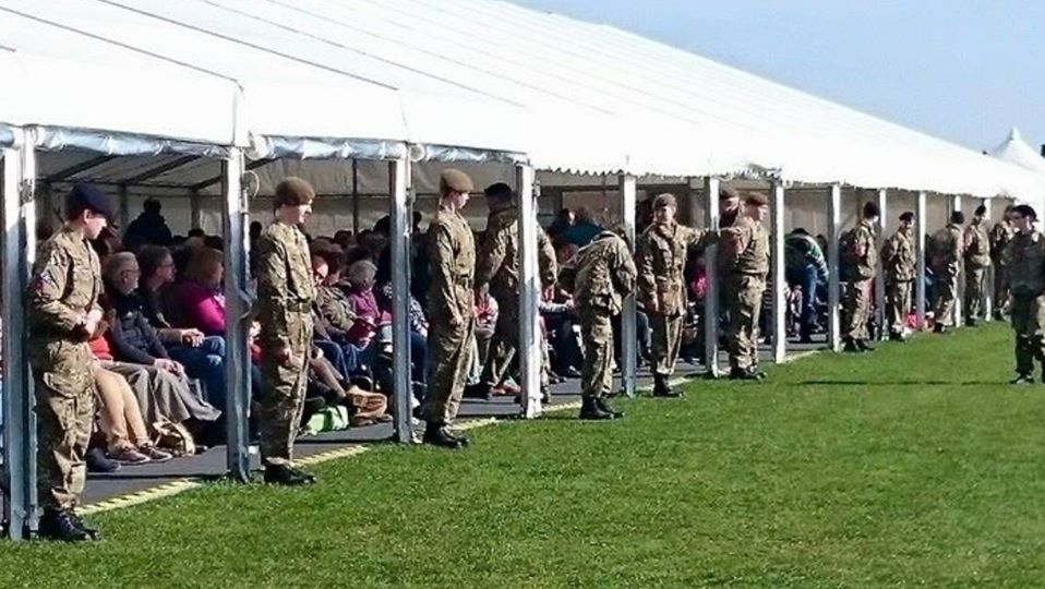 Soldiers in uniform stand in a line on grass, facing away from a white tent where people sit.