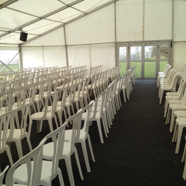 Rows of white plastic chairs in a tent, arranged for an event. Dark carpet, windows in the back.