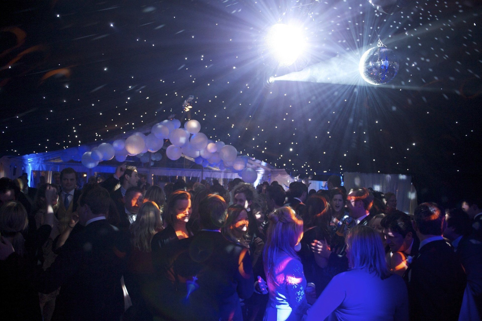 Disco ball illuminating a crowded dance floor with a tent-like ceiling and white balloons.