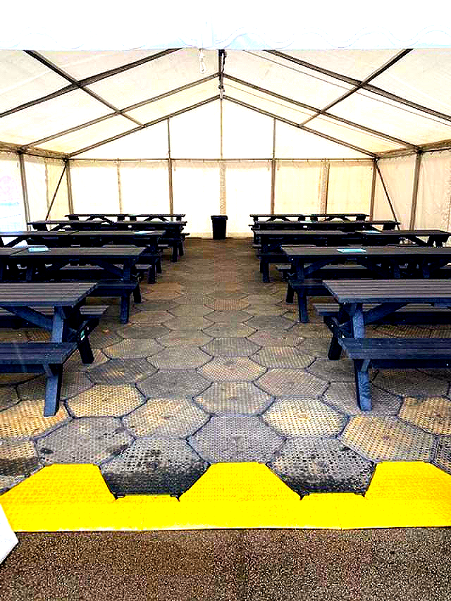 Inside tent with rows of picnic tables on hexagonal flooring. A yellow ramp is at the entrance.