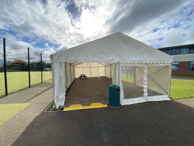 White tent set up on a schoolyard paved area, green trash can, and field in background.