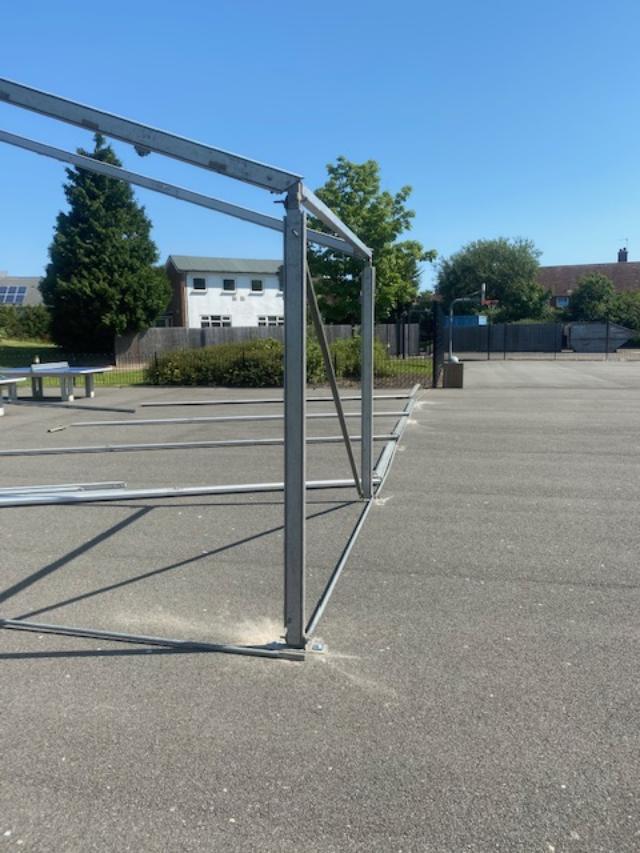 Metal frame structure on a gray paved area, likely an outdoor shelter. Blue sky.
