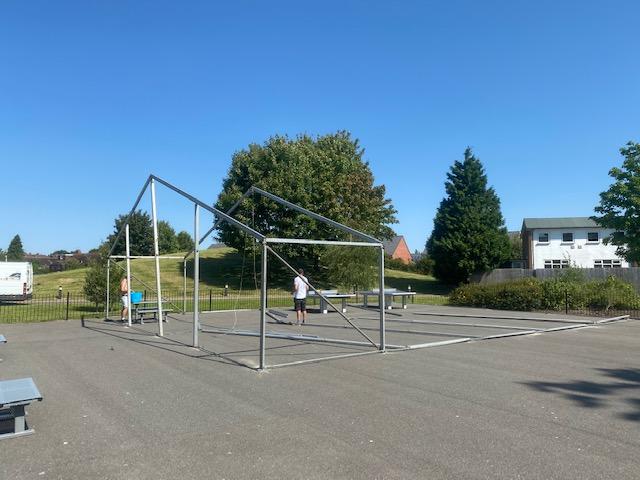 Two people assembling a metal frame tent on a paved surface on a sunny day.