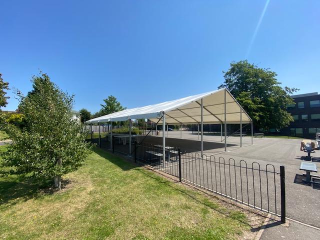 A long white tent on a sunny day in a grassy area with a black fence and a building in the background.