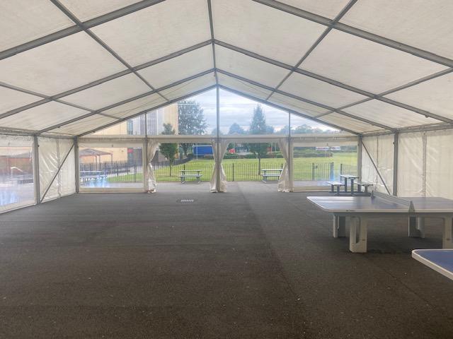 Inside a large, white-roofed tent with clear side panels, tables, and a view of a grassy area.