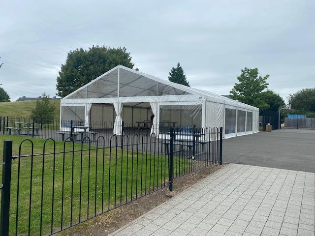 White tent with clear sides on a paved area, green grass and black fence in the foreground.