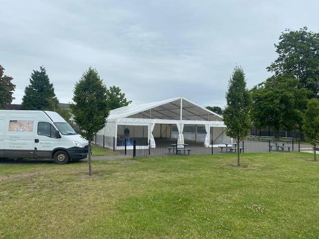 White tent set up outdoors, next to a van and some small trees on a green lawn.