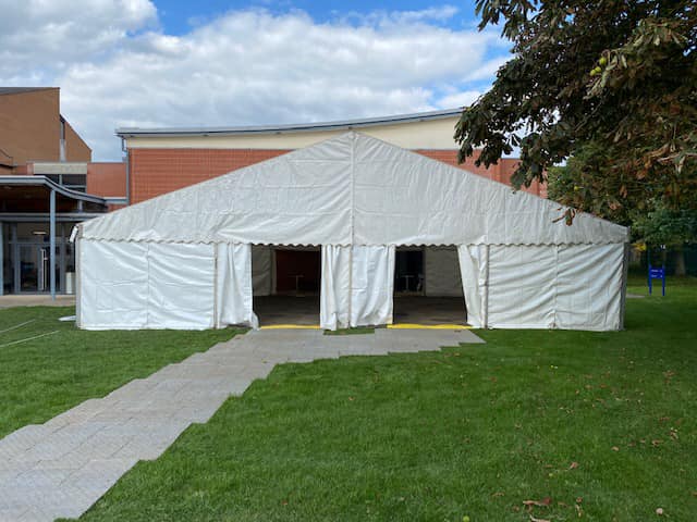 White tent on a green lawn with a gray path leading to the open entrances; a building is visible in the background.