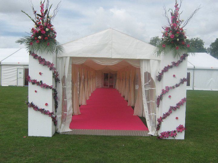 Entrance to a marquee, with pink carpet, floral decorations, and white pillars on green grass.