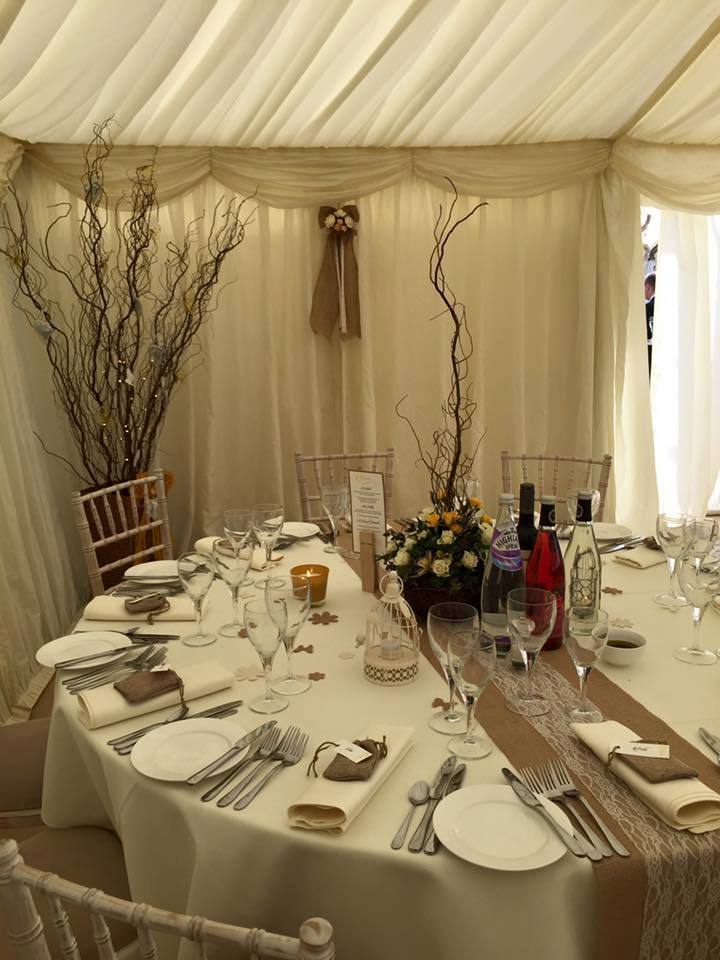 Wedding reception table with floral centerpieces, branches, and place settings inside a tent.