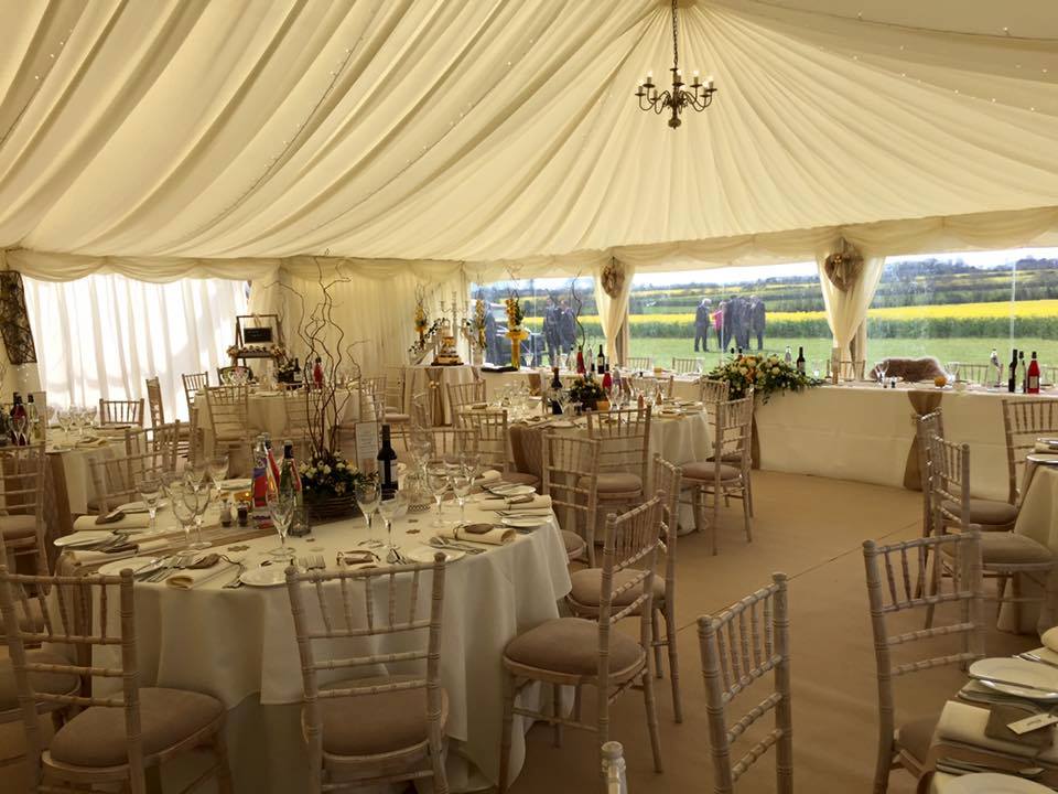 Inside a large tent set for a formal event. Round tables with white linens, chairs, and centerpieces.
